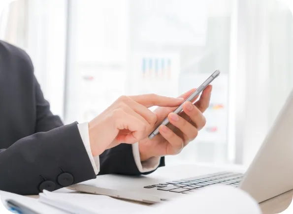 Hands of a person in a dark suit tapping a smartphone screen over a desk with an open laptop and paperwork, blurred charts visible in the background.