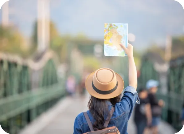 Rear view of a traveller in a tan wide-brimmed hat and denim jacket standing on a pedestrian bridge, holding a paper map aloft with the bridge and other people blurred in the background.