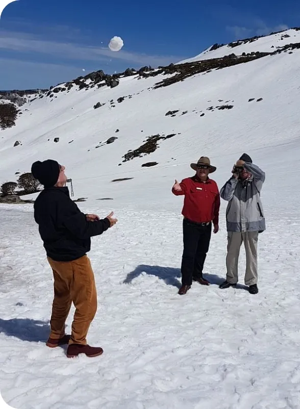 Three people in winter clothing stand on a snowy mountainside—one wearing a beanie prepares to catch a large snowball midair while a man in a red shirt gestures and a third person photographs the scene, with rocky snow-covered slopes and blue sky behind them.