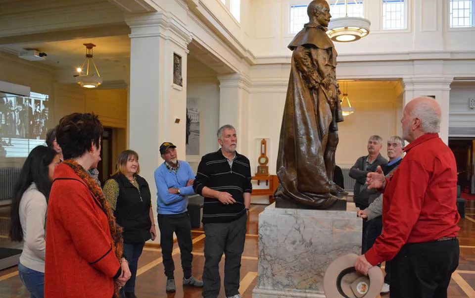 A group of visitors stand in a museum gallery around a tall bronze robed statue on a marble plinth as a man in a red shirt gestures while holding a hat, with high ceilings and classical columns in the background.