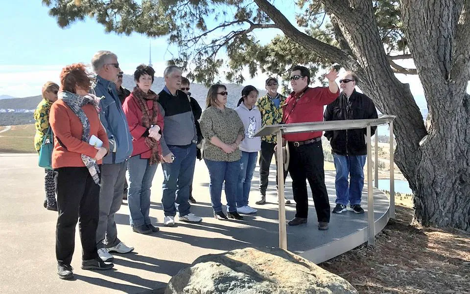 A tour guide in a red shirt gestures from a small metal lectern under a large tree while about a dozen people stand on a curved lookout platform listening, with rolling hills and a lake visible in the background.