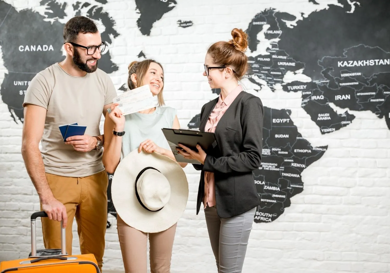 A smiling couple — the woman holding a wide-brimmed sun hat and boarding passes, the man holding passports and an orange suitcase — speak with a travel agent holding a clipboard in front of a large black-and-white world map painted on a brick wall.