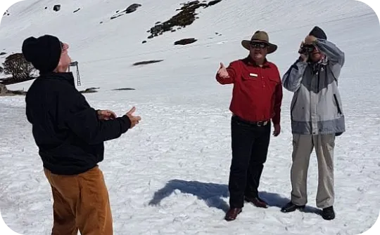 Three people stand on a snowy slope with scattered rocks: a person at left in a black beanie and brown pants gesturing with their hands, a central person in a red shirt and wide-brimmed hat giving a thumbs-up, and a person at right in a light jacket looking through binoculars or a camera.