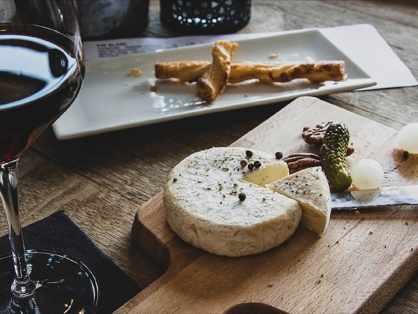 A cheese board with brie and pickles beside a glass of red wine.