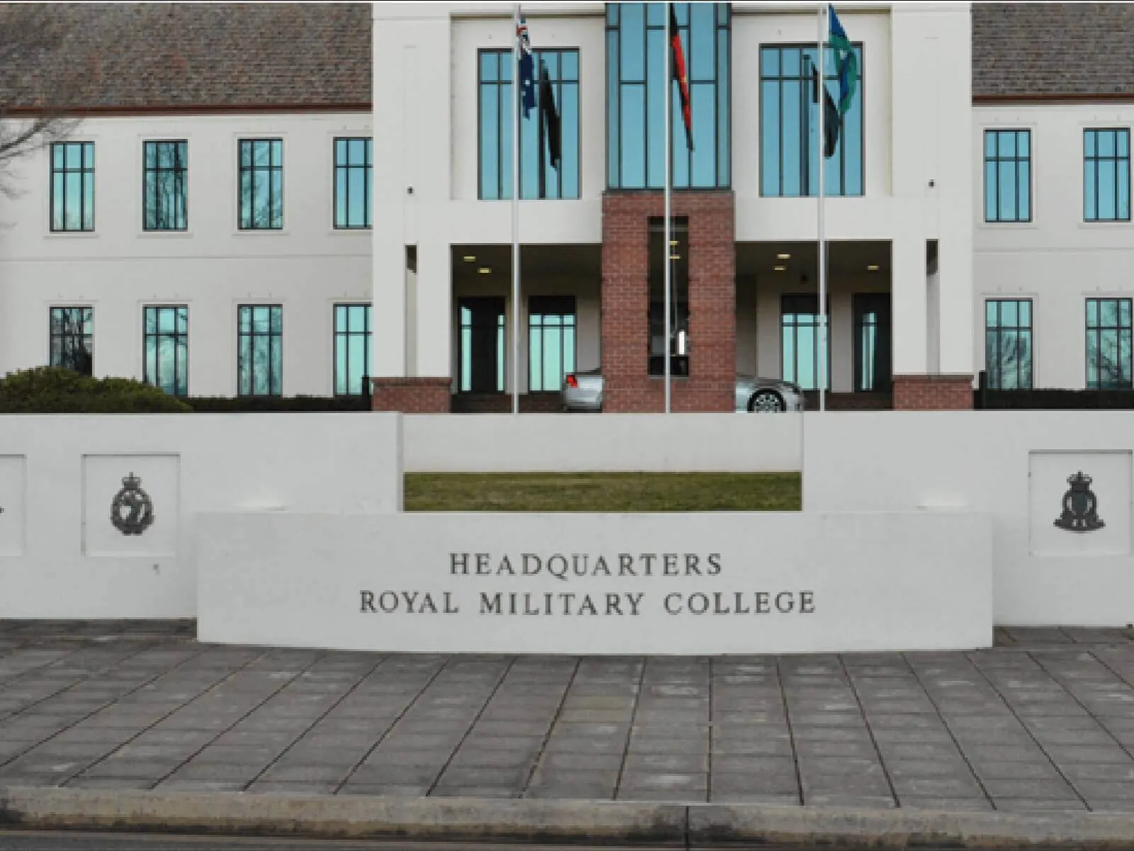 Front façade of the Headquarters Royal Military College showing a low white wall with engraved sign, a symmetrical white-and-brick building behind with tall windows, flagpoles and a car under the portico, and paved forecourt in the foreground.