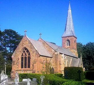 A sandstone church with a tall slate spire and a churchyard of headstones, under a blue sky.