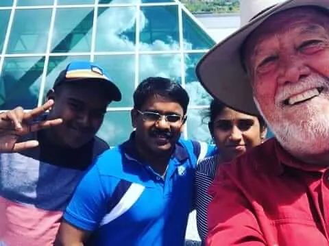 Four people smiling for a selfie in front of a glass‑paneled building: an older bearded man wearing a wide‑brimmed hat and red shirt at right, a man in a blue polo and glasses center, a person in a striped top behind him, and another man at left flashing a peace sign and wearing a cap.
