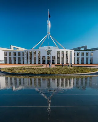 Parliament House in Canberra with its distinctive central flag mast and white colonnaded facade reflected in a foreground pool under a clear blue sky.