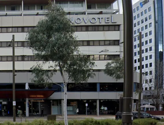 Novotel hotel façade across the street, framed by eucalyptus trees and tram lines, with pedestrians and parked cars along the urban sidewalk.
