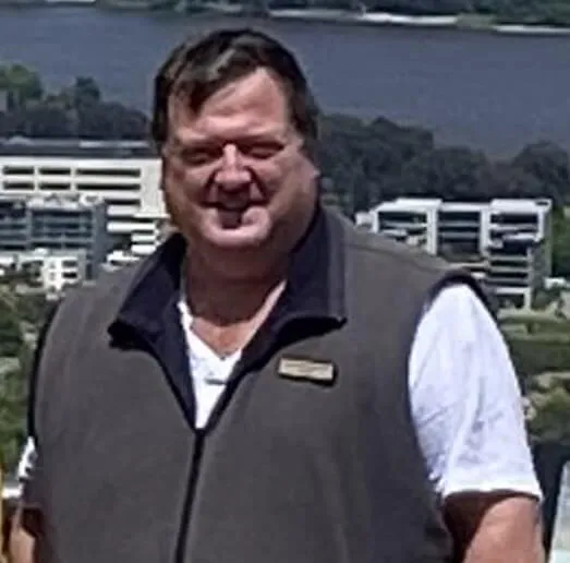 A smiling man wearing a dark grey zip-up vest over a white T-shirt, standing outdoors with a river and modern office buildings visible behind him.