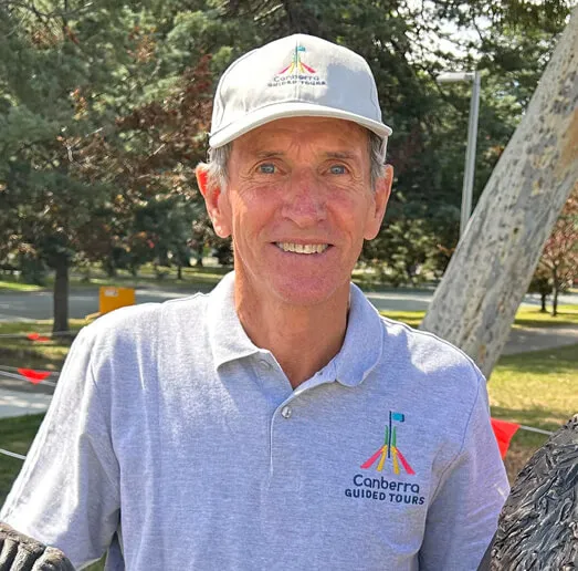 Smiling older man wearing a light grey polo shirt and matching cap embroidered with a colourful Canberra Guided Tours logo, standing outdoors in a park with trees and a pathway behind him.