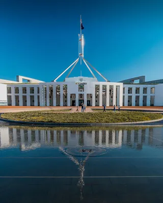 Parliament House in Canberra with its distinctive central flag mast and white colonnaded facade reflected in a foreground pool under a clear blue sky, with a few people on the forecourt.