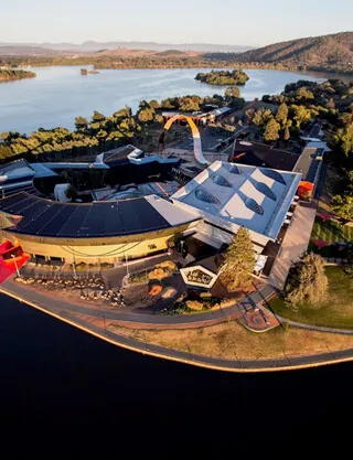 Aerial view of a modern lakeside entertainment complex with curved and peaked white roofs, a waterfront promenade and road at the water’s edge, surrounding trees and a lake stretching to tree-lined hills in the distance.