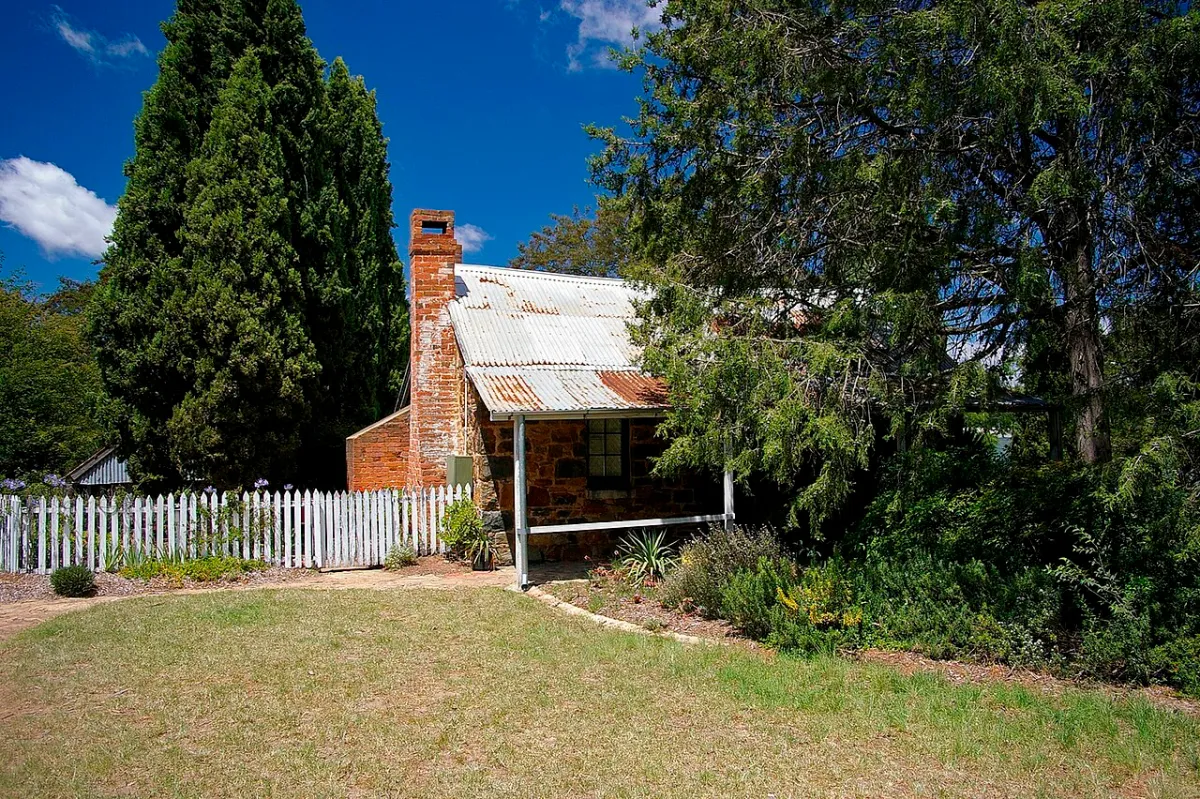 Blundells Cottage, a small stone cottage with a tin roof and a picket fence, among tall trees.