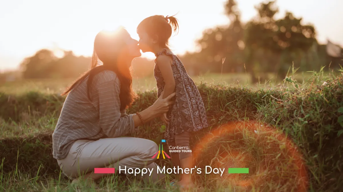 A mother kneeling in a grassy field at sunset leans forward to kiss her young daughter, warm backlighting and lens flare highlighting them with a 'Happy Mother's Day' greeting and Canberra Guided Tours logo overlaid at the bottom.