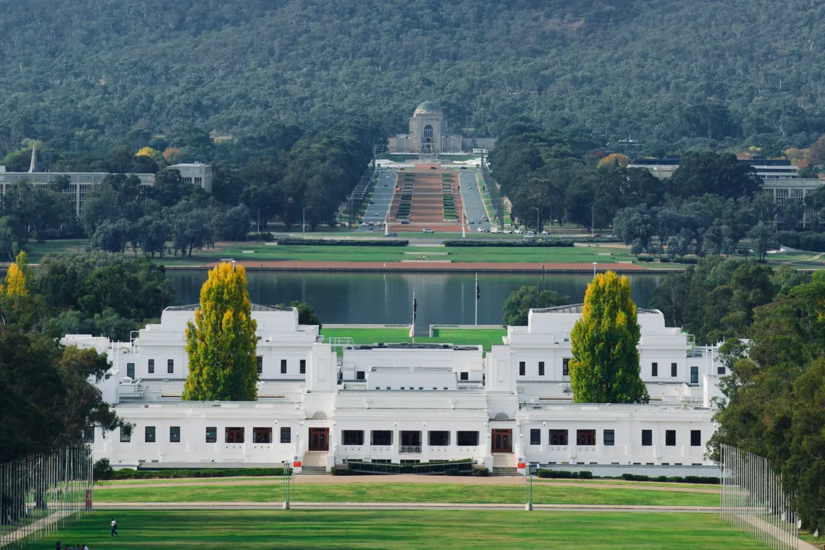White neoclassical government building fronting a wide green lawn and reflecting lake, with a tree-lined ceremonial avenue leading uphill to a domed stone memorial against a densely wooded hillside.