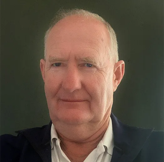 Head-and-shoulders portrait of an older man with short white hair and a slight smile, wearing a white collared shirt and dark jacket against a plain dark background.