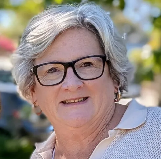 Smiling woman with short grey hair and black rectangular glasses, wearing a light collared top and small hoop earrings, photographed outdoors with blurred trees and sunlight in the background.