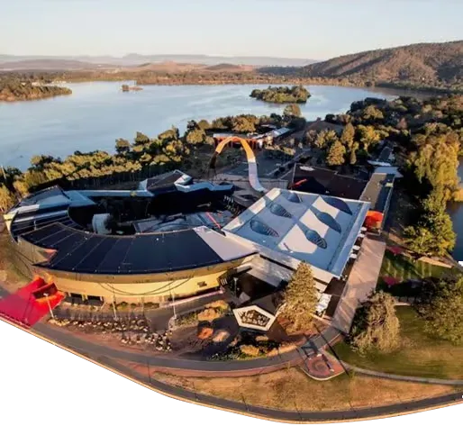Aerial view of a curved waterfront complex featuring a dark-roofed circular building and an adjoining white canopy with oval skylights, an orange arched ramp, and surrounding trees by a calm lake with islands and distant hills.