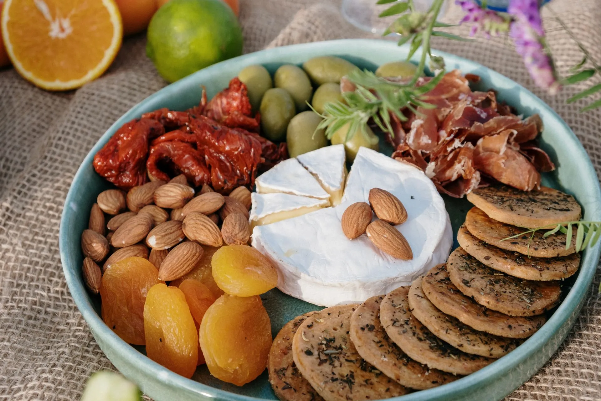A platter of brie, almonds, dried apricots, sun-dried tomatoes, olives, prosciutto and herb crackers on a hessian cloth.