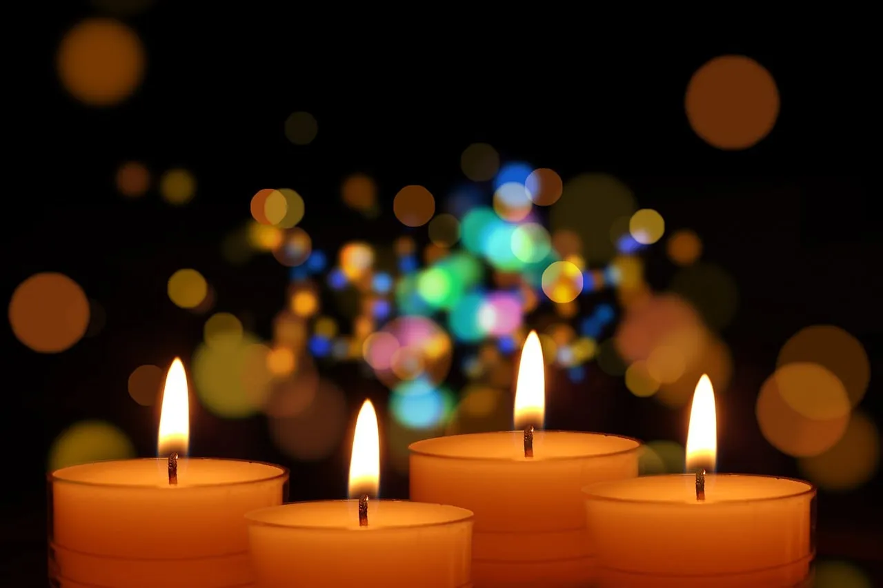 Four lit tealight candles in front of blurred, colourful lights.
